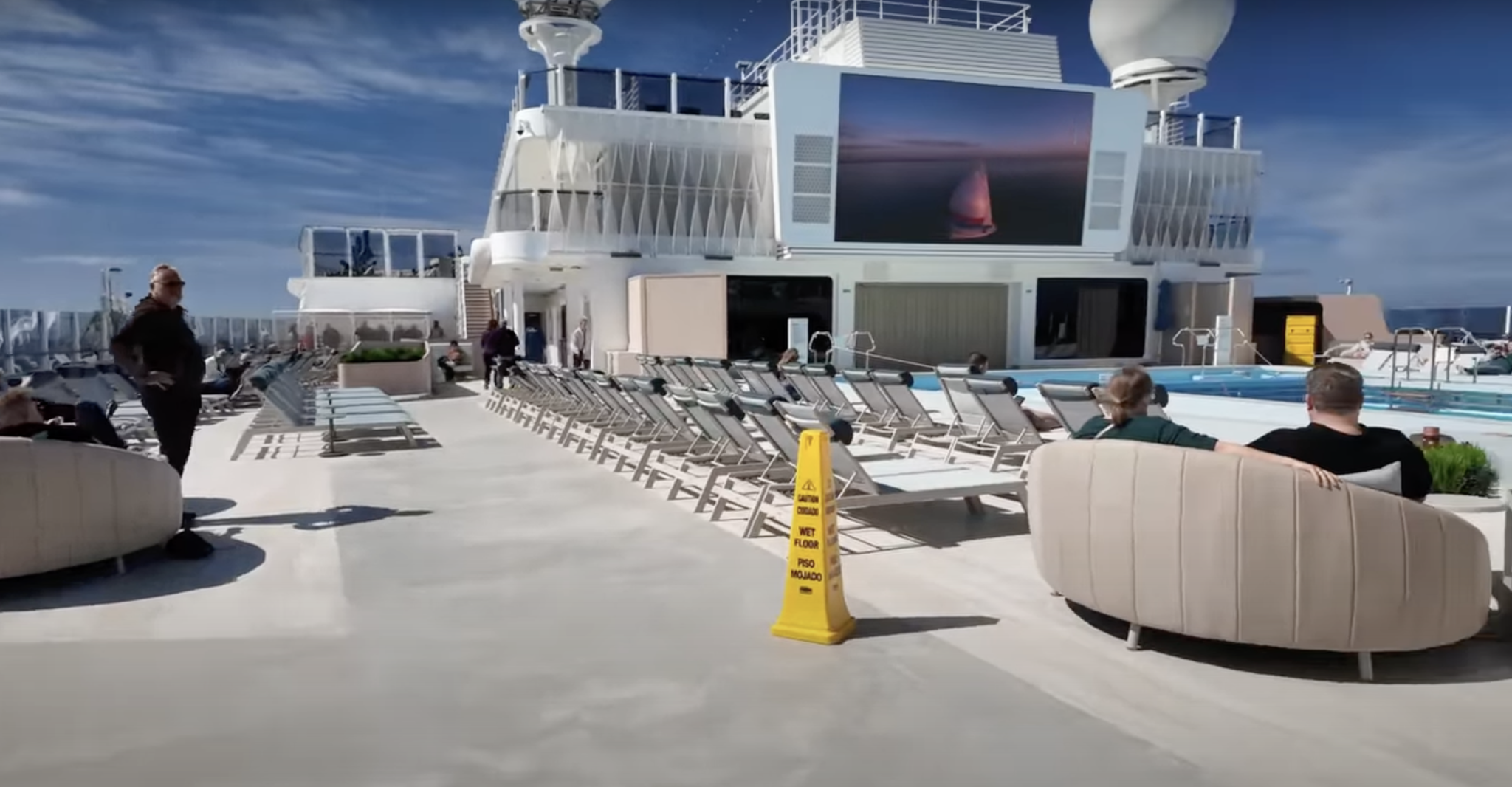 Wet floor caution sign on Norwegian Aqua pool deck, with lounge chairs and passengers in a sunny outdoor setting, highlighting potential slip and fall hazards related to cruise ship safety.